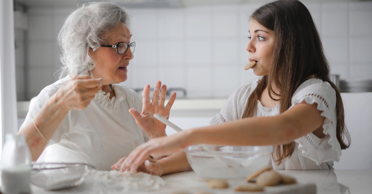 Femme âgée et adolescente calmes vêtues de vêtements décontractés se regardant et discutant tout en mangeant des cookies et préparant de la pâtisserie dans une cuisine moderne à la maison.