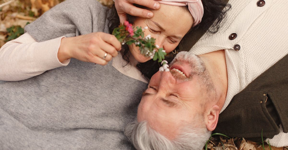 De dessus, la femme âgée rayonnante porte un grand foulard et un bandeau avec un bouquet de fleurs à la main, tandis que son mari plus âgé aux cheveux gris est allongé sur le sol, les yeux fermés, vêtu de vêtements chauds, parmi les feuilles tombées dans le parc.