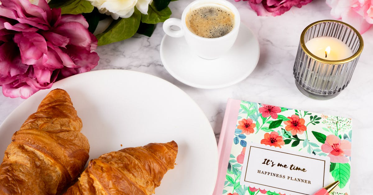 Disposition à plat d'un petit-déjeuner comprenant des croissants, du café, un agenda, une bougie et des fleurs.