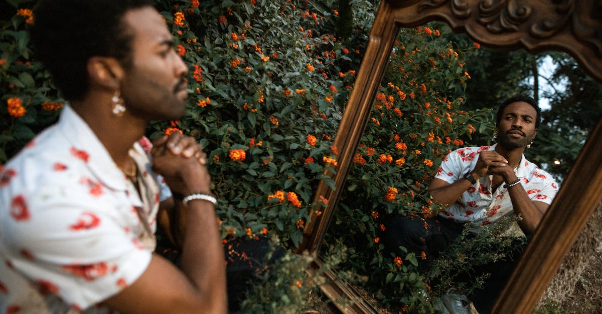 Un homme en chemise à motifs fleuris se reflète dans un miroir orné, entouré de fleurs éclatantes, arborant une expression pensive.